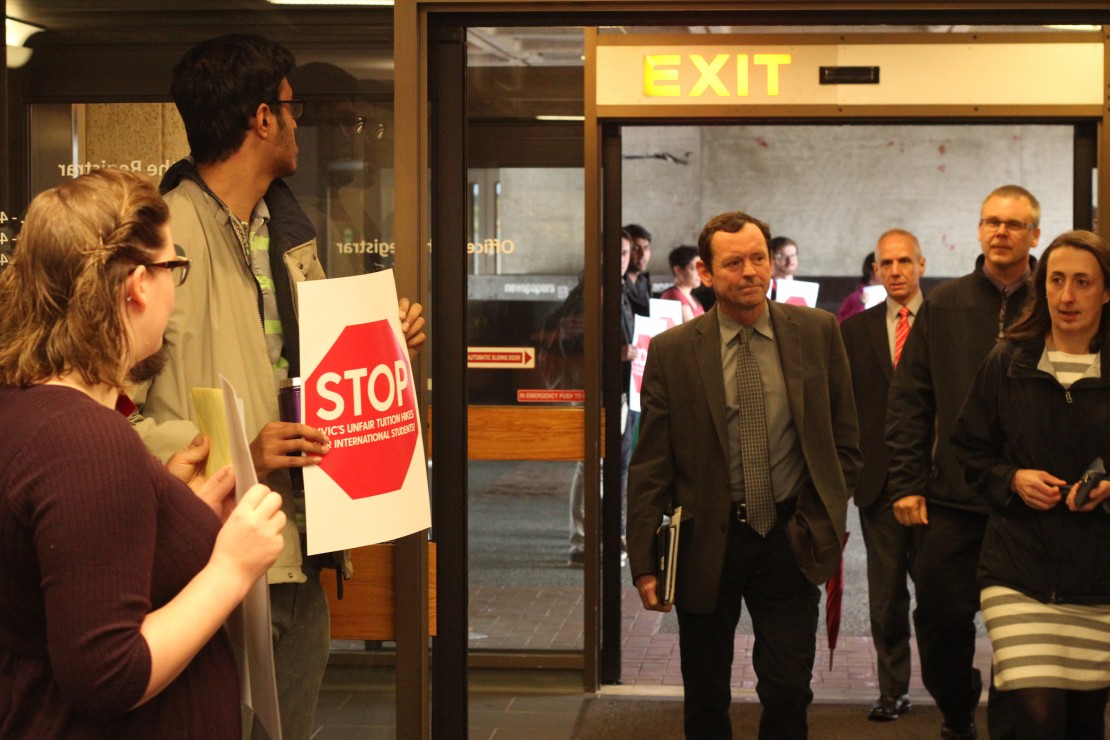 UVic President Jamie Cassels heads into the Board of Governors meeting Tuesday morning as protesters stand by with signs. The board voted in favour of implementing a four per cent increase to international student fees. Photo by Myles Sauer, Editor-in-Chief
