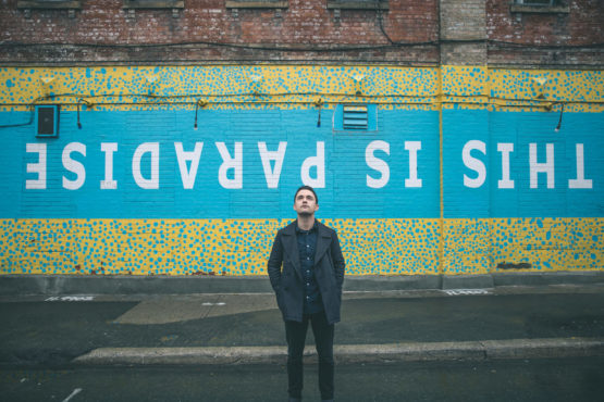 Joshua Hyslop stands in front of street art that says this is paradise. Hyslop released his latest folk album, Echoes.