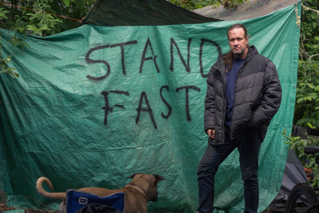 Shea Smith, a member of Victoria's street community, stands with his dog in front of a tarp that reads "stand fast"