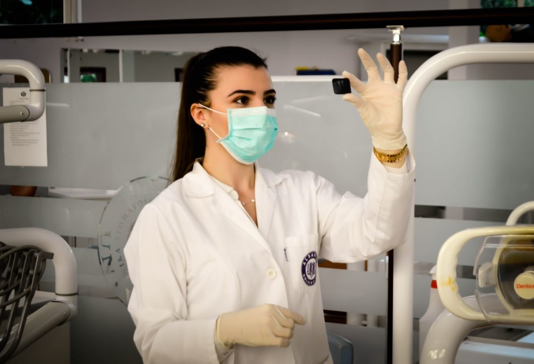 A woman in a medical coat, mask, and holding a needle in a hospital. Photo by Ani Kolleshi via Unsplash.